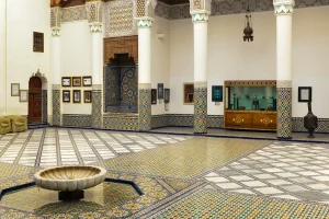 A courtyard in the Dar Si Said Museum, Marrakech, featuring colorful zellige tilework, carved wooden details, ornate columns, and a central marble fountain.