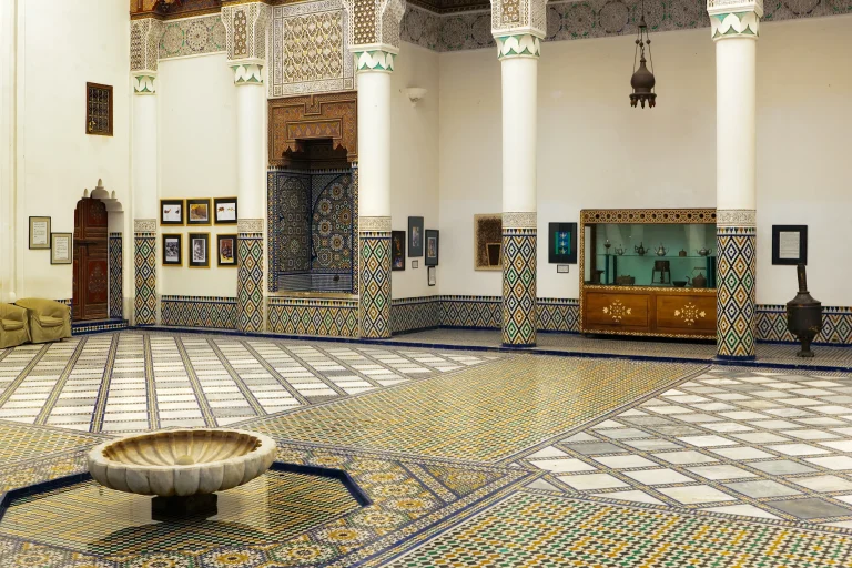 A courtyard in the Dar Si Said Museum, Marrakech, featuring colorful zellige tilework, carved wooden details, ornate columns, and a central marble fountain.