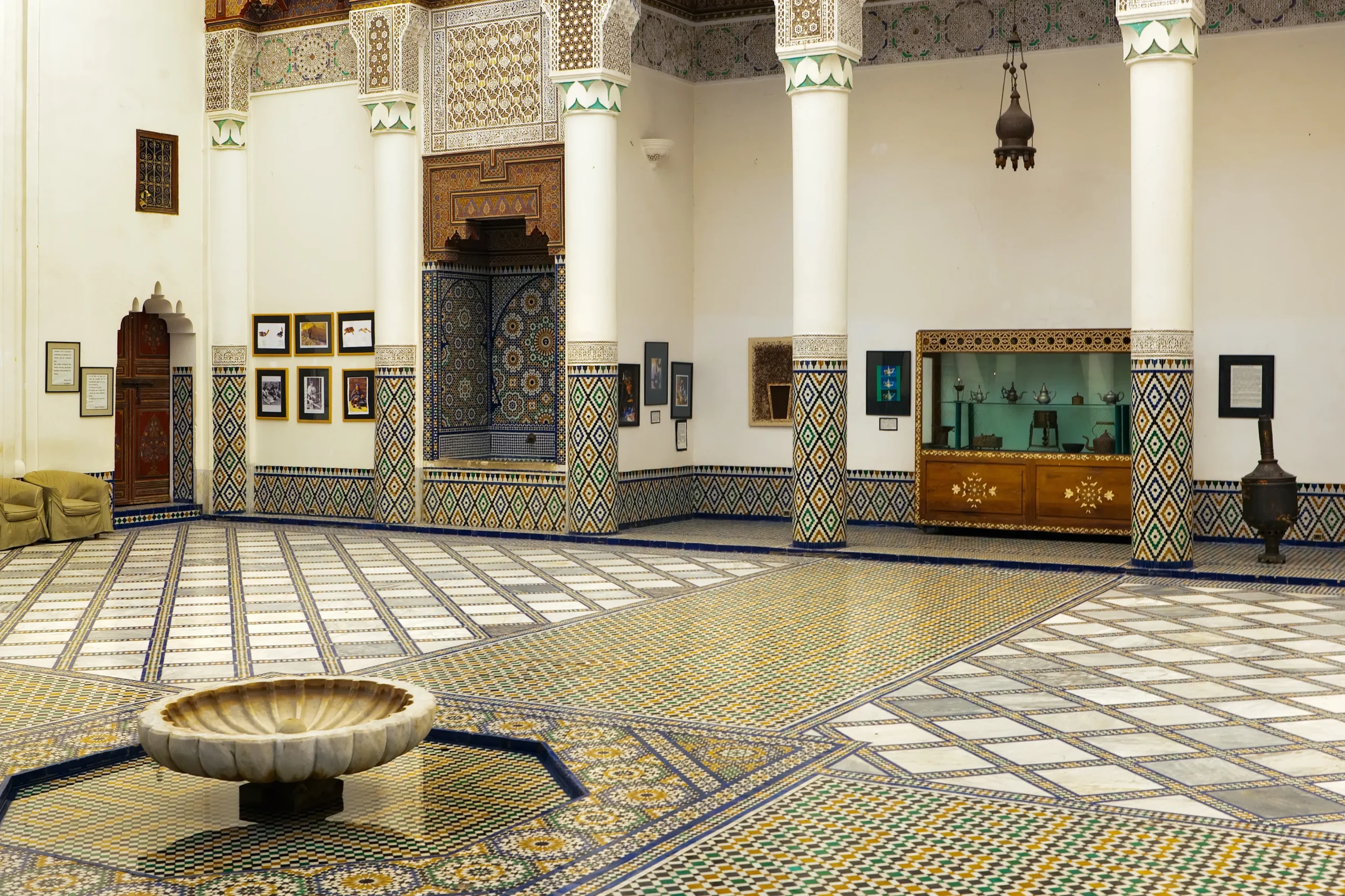 A courtyard in the Dar Si Said Museum, Marrakech, featuring colorful zellige tilework, carved wooden details, ornate columns, and a central marble fountain.