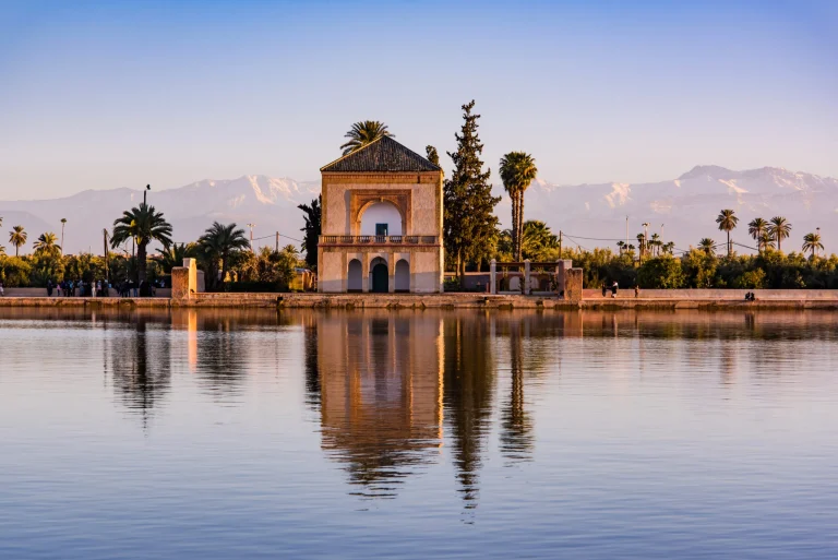 A view of the Menara Gardens in Marrakech, featuring a historic pavilion beside a reflective pool, framed by olive groves, with the Atlas Mountains in the background.
